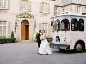 Newport Rhode Island Wedding Photographers | Deirdre Photography Timeless and Organic Moments | A newlywed couple kisses in front of a grand mansion. The bride in a flowing white dress holds a bouquet, and a vintage white trolley is parked nearby.