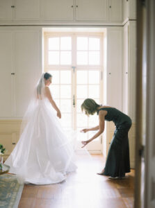 Bride standing beside tall windows while a bridesmaid adjusts her gown in soft natural light before the celebration at Glen Manor House.
