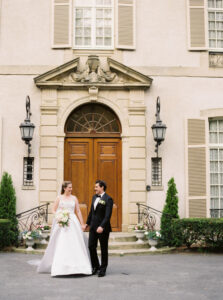 Newport Rhode Island Wedding Photographers | Deirdre Photography Timeless and Organic Moments | A bride in a white gown and a groom in a black tuxedo stand holding hands, smiling, in front of a grand building with ornate doors and elegant lanterns.
