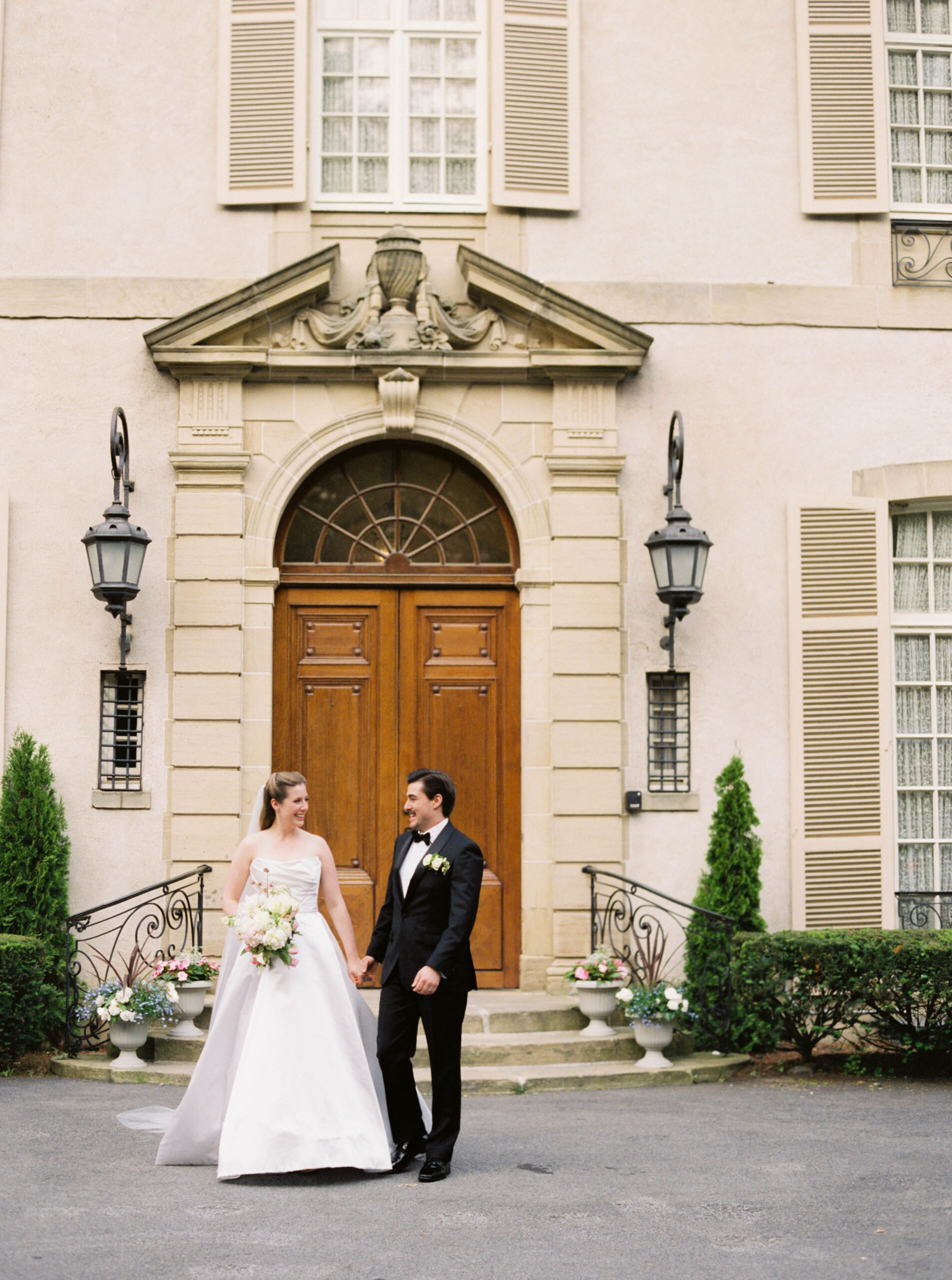 Newport Rhode Island Wedding Photographers | Deirdre Photography Timeless and Organic Moments | A bride in a white gown and a groom in a black tuxedo stand holding hands, smiling, in front of a grand building with ornate doors and elegant lanterns.