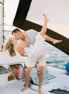 Wedding Photographer Newport Rhode Island | Deirdre Photography Timeless and Organic Moments | A couple shares a joyful kiss on a sailboat, with the man lifting the woman playfully. The sea and sails frame the scene