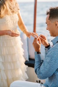 Engagement Photos Rhode Island | Deirdre Photography Timeless and Organic Moments | A man in a blue shirt kneels, placing a ring on a woman's finger. She wears a ruffled white dress. They are on a boat, with the ocean in the background. Romantic atmosphere.