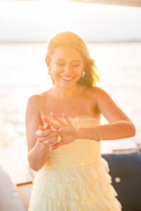 Engagement Photos Rhode Island | Deirdre Photography Timeless and Organic Moments | A woman in a light yellow dress stands on a boat, smiling as she applies lotion to her hands. The sun sets behind her, emitting a warm, golden glow.