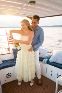 Engagement Photos Rhode Island | Deirdre Photography Timeless and Organic Moments | A joyful couple on a boat at sunset, with the woman in a flowing yellow dress opening a champagne bottle, and the man smiling behind her.