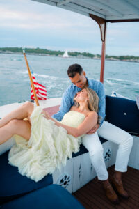 Engagement Photos Rhode Island | Deirdre Photography Timeless and Organic Moments | Couple on a boat, woman in a yellow dress leaning against a man in a blue shirt. They share a joyful moment, background shows water and an American flag.