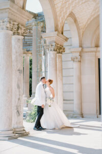 Castle Hill Wedding | Deirdre Photography Timeless and Organic Moments | A bride and groom share a kiss under elegant, sunlit stone arches. The bride holds a bouquet, while both radiate joy and elegance in formal attire.