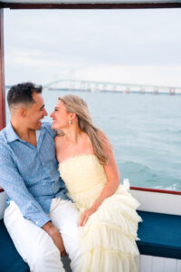 Engagement Photos Rhode Island | Deirdre Photography Timeless and Organic Moments | A couple sits closely, smiling at each other on a boat with a scenic bridge and calm water in the background.