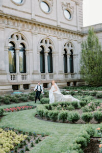 Castle Hill Wedding | Deirdre Photography Timeless and Organic Moments | A bride and groom joyfully run through a manicured garden in front of an ornate building with large arched windows