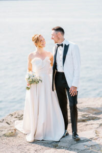Castle Hill Wedding | Deirdre Photography Timeless and Organic Moments | A couple in wedding attire stands smiling on a rocky shoreline. The bride wears a white gown and holds a bouquet, while the groom wears a white tuxedo.