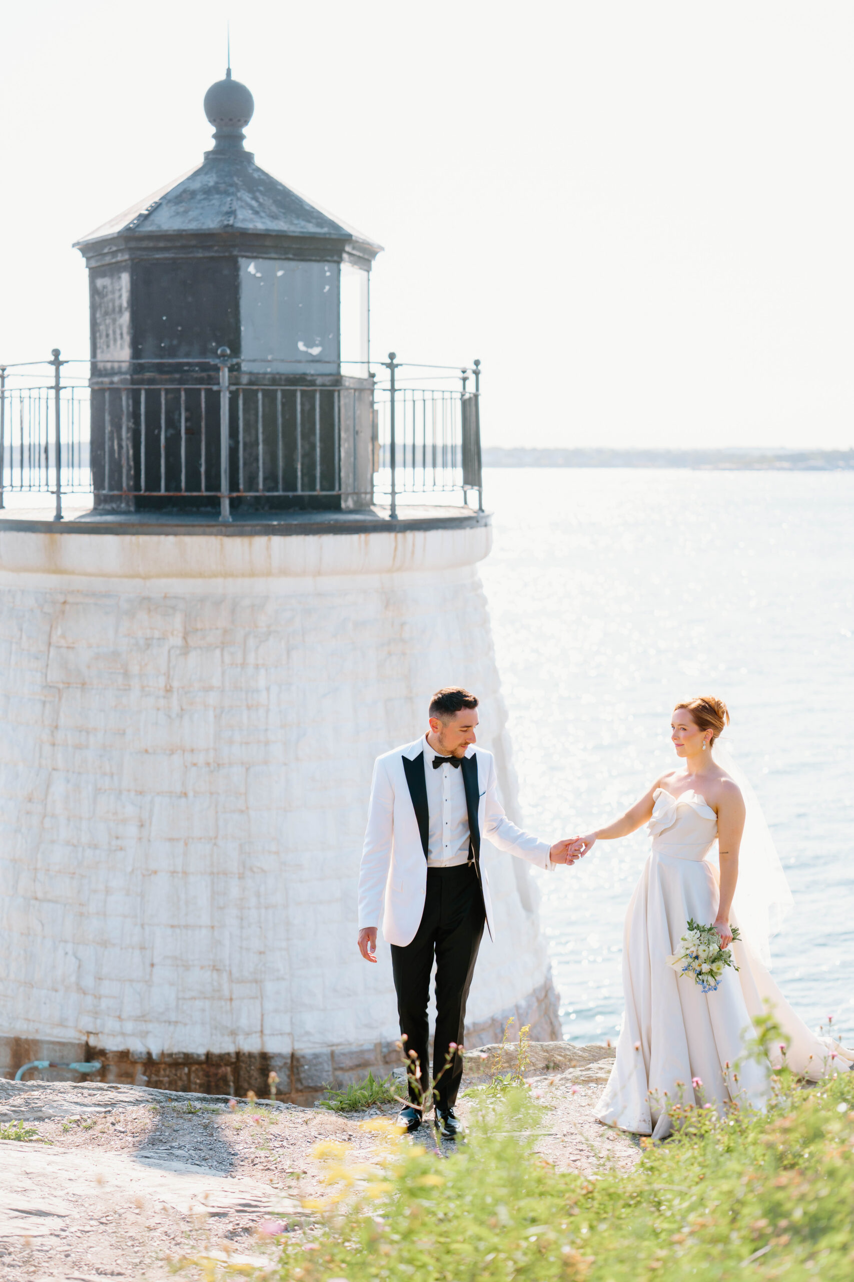 Castle Hill Wedding | Deirdre Photography Timeless and Organic Moments | Bride in white gown and groom in white tuxedo hold hands by a lighthouse overlooking a sparkling sea.