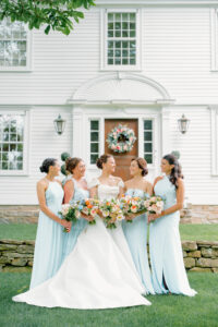 Smith Farm Gardens Wedding | Deirdre Photography Timeless and Organic Moments | A bride in a white gown stands with four bridesmaids in light blue dresses, all holding colorful bouquets, smiling in front of a white house.