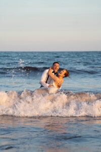 Castle Hill Wedding | Deirdre Photography Timeless and Organic Moments | A joyful couple in formal attire playfully embrace in ocean waves at sunset, radiating happiness and love, with water splashing around them.