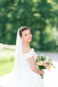 The bride turns back over her shoulder with her bouquet in hand, framed by soft greenery in a luminous portrait by a fine art wedding photographer in Rhode Island.