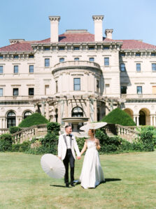 Castle Hill Wedding | Deirdre Photography Timeless and Organic Moments | A couple holds hands in front of a grand, historic mansion. They smile, holding white parasols, exuding elegance and joy against the clear blue sky.