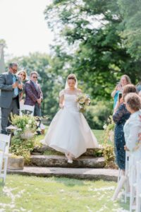Smith Farm Gardens Wedding | Deirdre Photography Timeless and Organic Moments | A bride in an off-shoulder white dress holds a bouquet, descending stone steps surrounded by guests. The outdoor setting is lush and green.