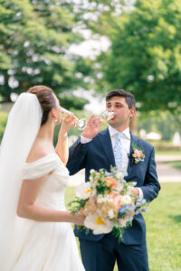 Smith Farm Gardens Wedding | Deirdre Photography Timeless and Organic Moments | A bride and groom stand outdoors, toasting with champagne. The bride holds a colorful bouquet, and the groom wears a blue suit with a floral boutonniere.