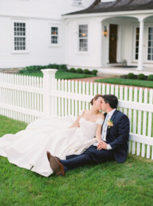 Smith Farm Gardens Wedding | Deirdre Photography Timeless and Organic Moments | A bride and groom sit on grass, kissing gently beside a white picket fence. The bride wears an elegant white dress, while the groom is in a navy suit with a boutonniere. The scene is serene and romantic, set in front of a classic white house.
