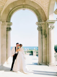 Beneath a grand stone arch, the couple pauses in the bright coastal light with the ocean beyond, a timeless scene often sought from a Newport RI wedding photographer.