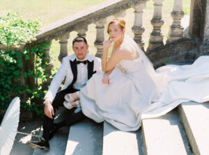 Wedding Photographer in Rhode Island | Deirdre Photography Timeless and Organic Moments | A bride and groom sit on stone steps, the bride in a flowing white gown and veil, and the groom in a black and white tuxedo. They appear relaxed and content, with a lush garden in the background.