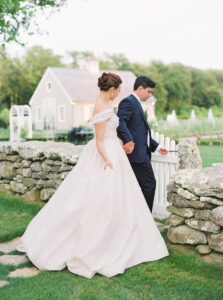 Smith Farm Gardens Wedding | Deirdre Photography Timeless and Organic Moments | Bride and groom holding hands, walking through a stone archway in a lush garden. Bride in an off-shoulder gown, groom in a navy suit, quaint cottage in background.