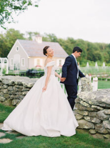 Smith Farm Gardens Wedding | Deirdre Photography Timeless and Organic Moments | A bride in an elegant white gown smiles joyfully while holding hands with a groom in a navy suit. They stand by a rustic stone wall, with a quaint cottage and lush greenery in the background.