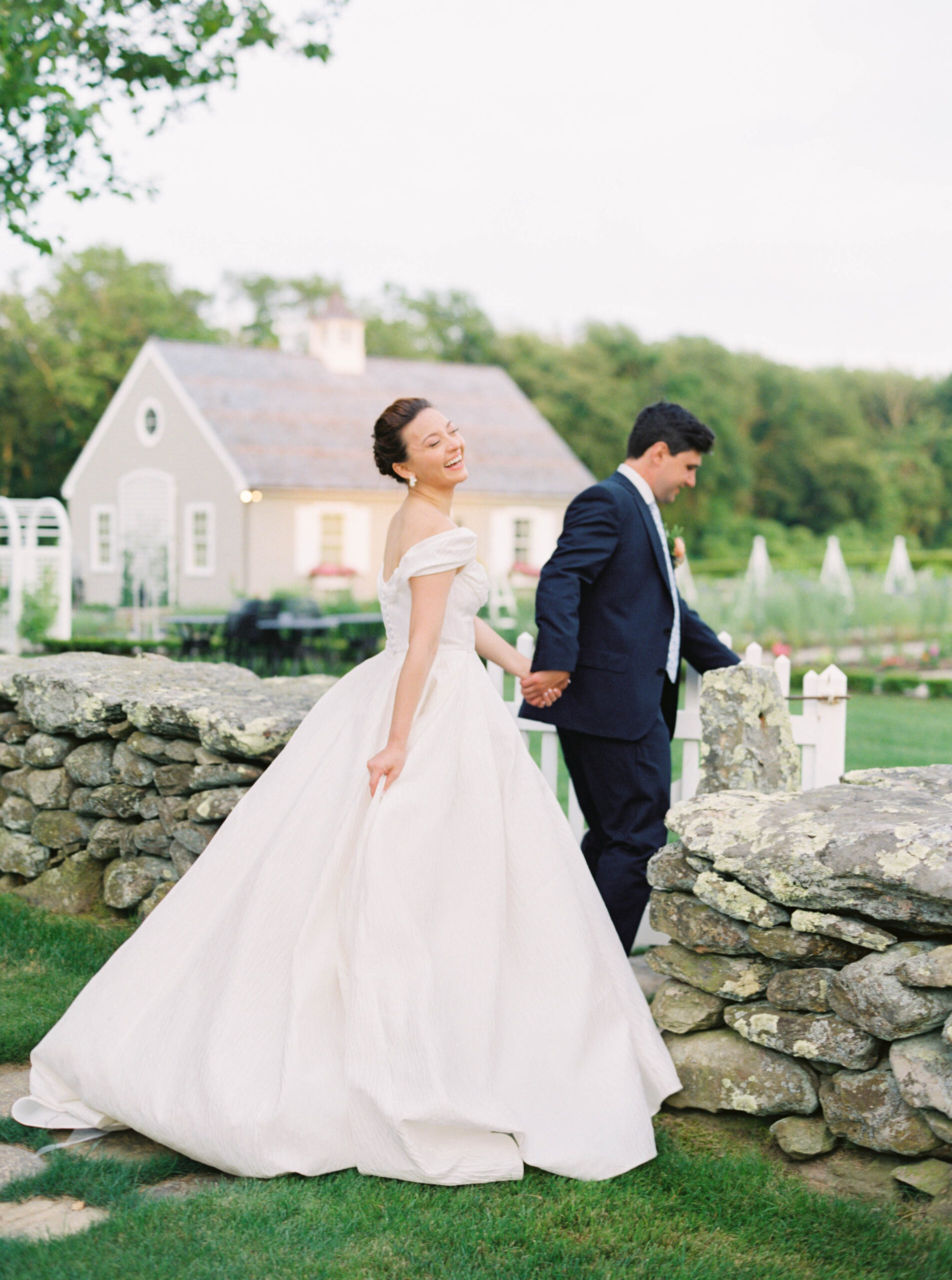 Smith Farm Gardens Wedding | Deirdre Photography Timeless and Organic Moments | A bride in an elegant white gown smiles joyfully while holding hands with a groom in a navy suit. They stand by a rustic stone wall, with a quaint cottage and lush greenery in the background.