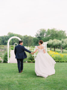 Smith Farm Gardens Wedding | Deirdre Photography Timeless and Organic Moments | A couple in wedding attire walks hand in hand through a lush garden, beneath a white arch