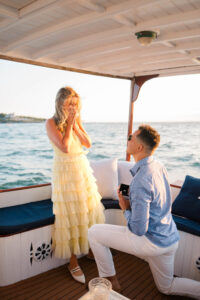 Engagement Photos Rhode Island | Deirdre Photography Timeless and Organic Moments | A man kneels on a boat, proposing with a ring to a surprised woman in a yellow dress. They are surrounded by ocean under a clear blue sky.