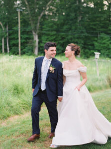 Smith Farm Gardens Wedding | Deirdre Photography Timeless and Organic Moments | A couple walks hand in hand through a lush, green field. The groom wears a navy suit and floral tie, while the bride is in a flowing white gown. They look at each other, smiling warmly.