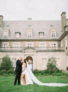 Aldrich Mansion Wedding Photos | Deirdre Photography Timeless and Organic Moments | Bride and groom share a tender kiss in front of a grand, stone mansion with manicured gardens. Her long veil flows elegantly on the grass.