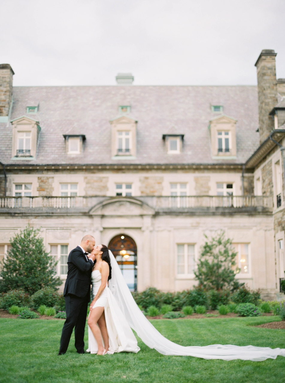 Aldrich Mansion Wedding Photos | Deirdre Photography Timeless and Organic Moments | Bride and groom share a tender kiss in front of a grand, stone mansion with manicured gardens. Her long veil flows elegantly on the grass.