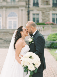 Aldrich Mansion Wedding Photos | Deirdre Photography Timeless and Organic Moments | A joyful couple in wedding attire shares a tender moment outside an elegant stone building. The bride holds a bouquet of white flowers.