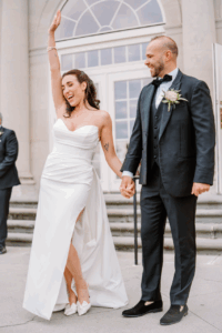 Aldrich Mansion Wedding Photos | Deirdre Photography Timeless and Organic Moments | A joyful bride in a white gown with a thigh-high slit and groom in a black suit stand hand-in-hand, celebrating outside a building with arched windows.