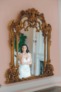 A bride is captured in an ornate gold mirror, her reflection softly lit in a pastel-toned room that highlights the romantic details of Newport Rhode Island wedding venues.