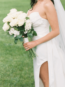 The Chanler at Cliff Walk Wedding | Deirdre Photography Timeless and Organic Moments | A bride in a white strapless gown holds a bouquet of white roses. The setting is an outdoor grassy area, conveying a serene and joyful wedding scene.