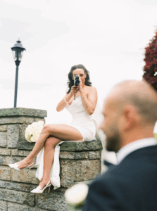 Seated on a stone wall, the bride playfully points a camera toward the groom in a candid moment that reflects the creative eye of photographers in RI.