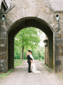 Newport RI Mansion Wedding | Deirdre Photography Timeless and Organic Moments | A bride and groom share a kiss under a stone archway, surrounded by lush greenery. The scene is romantic and serene, capturing a tender moment.