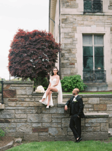 Aldrich Mansion Wedding Photos | Deirdre Photography Timeless and Organic Moments | A woman in a white dress sits on a stone wall, while a man in a suit stands below, looking up at her. They are beside a stone building and a red-leaved tree.