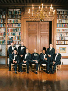 Aldrich Mansion Wedding Photos | Deirdre Photography Timeless and Organic Moments | A group of eight men in formal black suits and bow ties pose in a grand library with wooden shelves filled with books, under an elegant chandelier.