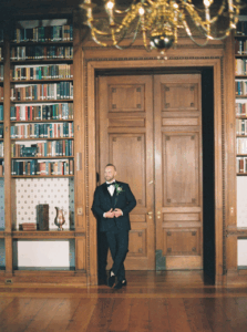 Rhode Island Wedding Photographer | Deirdre Photography Timeless and Organic Moments | Man in a dark suit stands relaxed against an ornate wooden door in a library, surrounded by shelves of colorful books. Warm, elegant atmosphere.