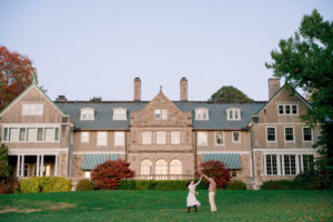 Blithewold Mansion, Gardens and Arboretum | Deirdre Photography Timeless and Organic Moments | A couple dances joyfully on a lush green lawn in front of a large, elegant stone mansion under a clear sky, conveying a sense of romance and tranquility.