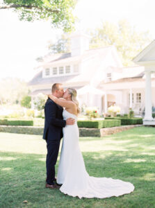 Smith Farm Garden Wedding | Deirdre Photography Timeless and Organic Moments | A bride in a white gown and a groom in a dark suit kiss on a sunlit lawn in front of a charming house, conveying love and joy on their wedding day.