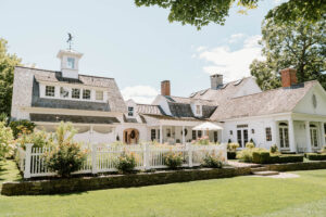 Newport RI Mansion Wedding | Deirdre Photography Timeless and Organic Moments | Charming white colonial house with gabled roofs, a white picket fence, lush greenery, and a clear blue sky, exuding a tranquil and welcoming vibe.
