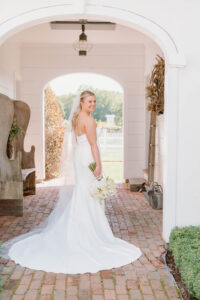 Smith Farm Garden Wedding | Deirdre Photography Timeless and Organic Moments | A bride in a white gown stands on a brick path under an archway, holding a bouquet. Sunlight filters in, creating a serene and elegant atmosphere.
