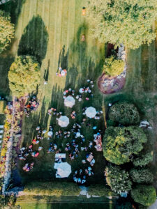 Smith Farm Garden Wedding | Deirdre Photography Timeless and Organic Moments | Aerial view of a garden party with scattered groups sitting on grass, surrounded by trees. White umbrellas provide shade, creating a festive atmosphere.