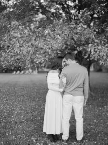 Blithewold Mansion, Gardens and Arboretum | Deirdre Photography Timeless and Organic Moments | A couple stands closely in a serene park, facing away, beneath a large tree. The monochrome setting conveys a romantic, timeless mood.