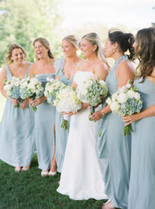 Smith Farm Garden Wedding | Deirdre Photography Timeless and Organic Moments | A bride in a white dress stands with bridesmaids in light blue dresses, all holding bouquets. They smile joyfully outdoors on a sunny day.