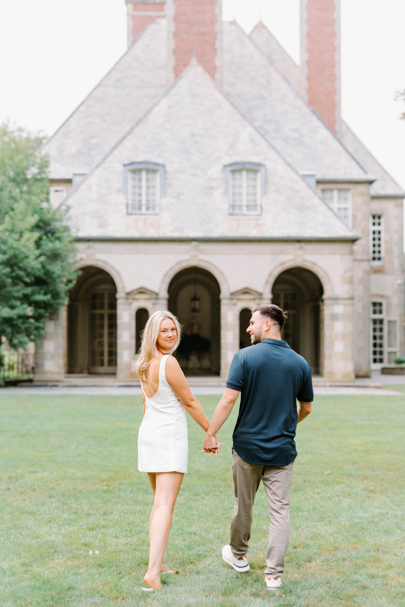 Glen Manor House Portsmouth RI | Deirdre Photography Timeless and Organic Moments | A couple holding hands walks toward a large, elegant stone house with arched doorways. The woman smiles back at the camera, creating a joyful tone.