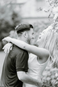Glen Manor House Portsmouth RI | Deirdre Photography Timeless and Organic Moments | A black and white photo of a couple embracing and laughing joyfully outdoors. The scene conveys affection and happiness, surrounded by blurred greenery.