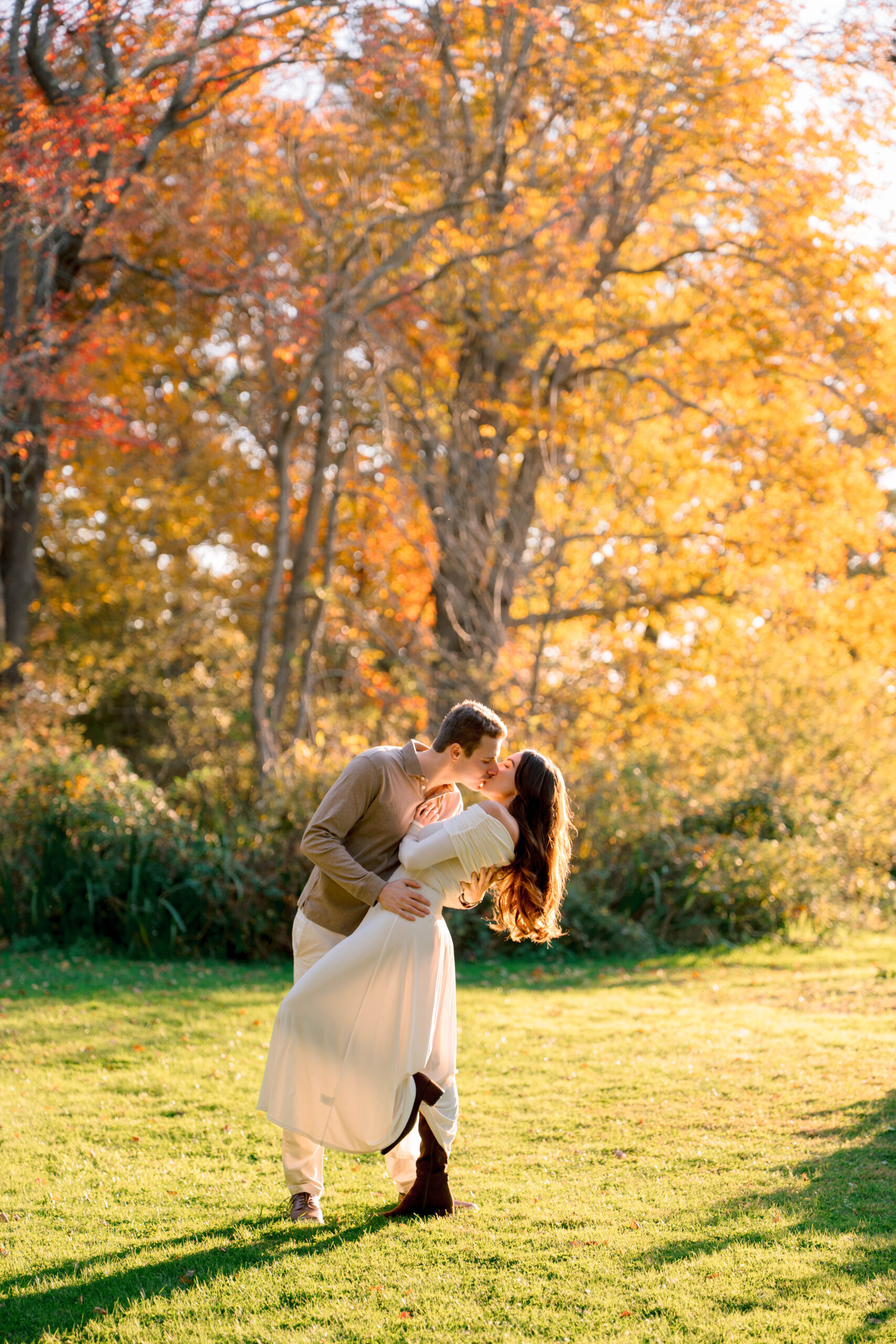 Aldrich Mansion Wedding Photos | Deirdre Photography Timeless and Organic Moments | A couple embraces romantically under vibrant orange autumn trees in a sunlit park. The woman's white dress contrasts with the lush green grass.